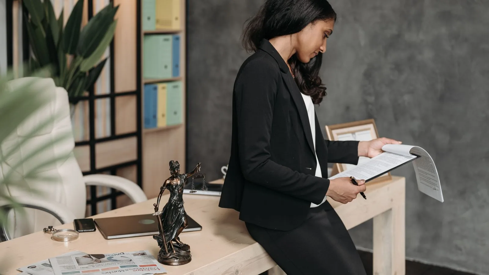 A lawyer in a black suit reviews a legal document while leaning against her office desk, a Lady Justice statuette beside her.