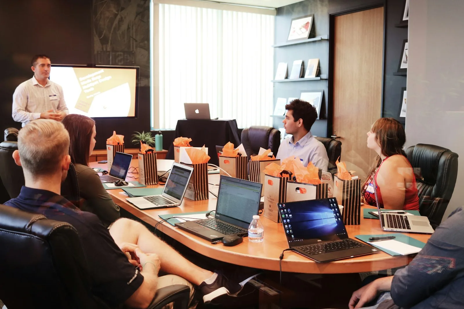 A consultant presenting to a team in a glass-walled boardroom with laptops open