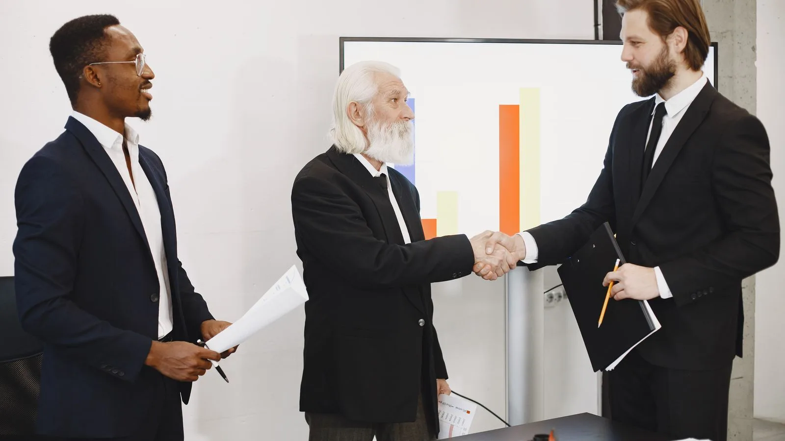 Three businessmen in suits having a meeting, shaking hands in front of a presentation chart.