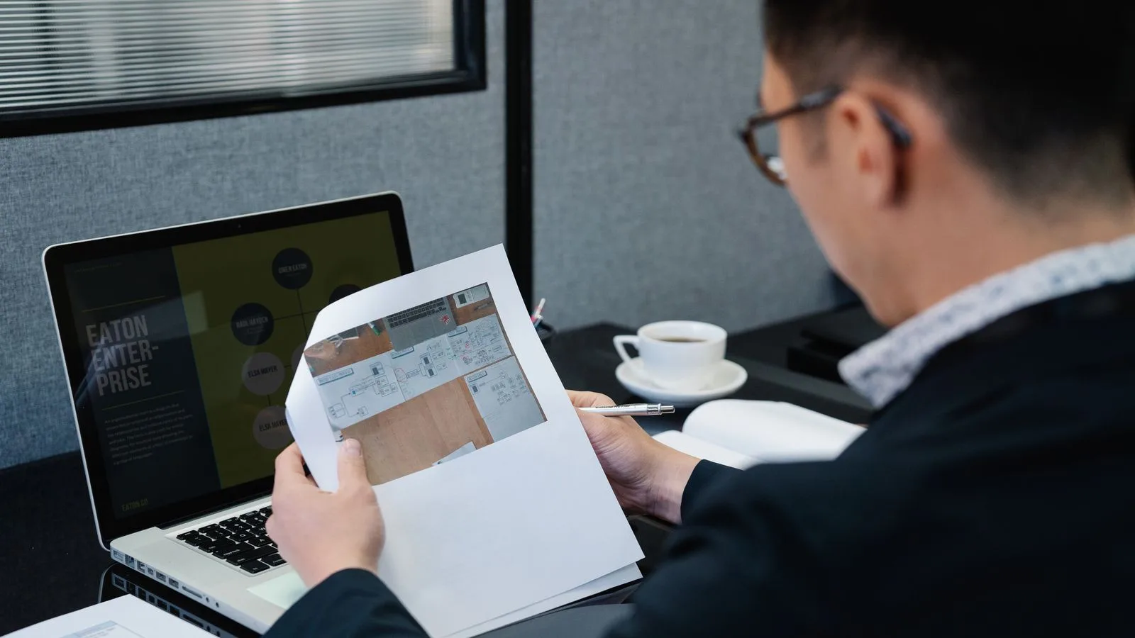 A man in an office setting analyzing documents with a laptop open nearby.