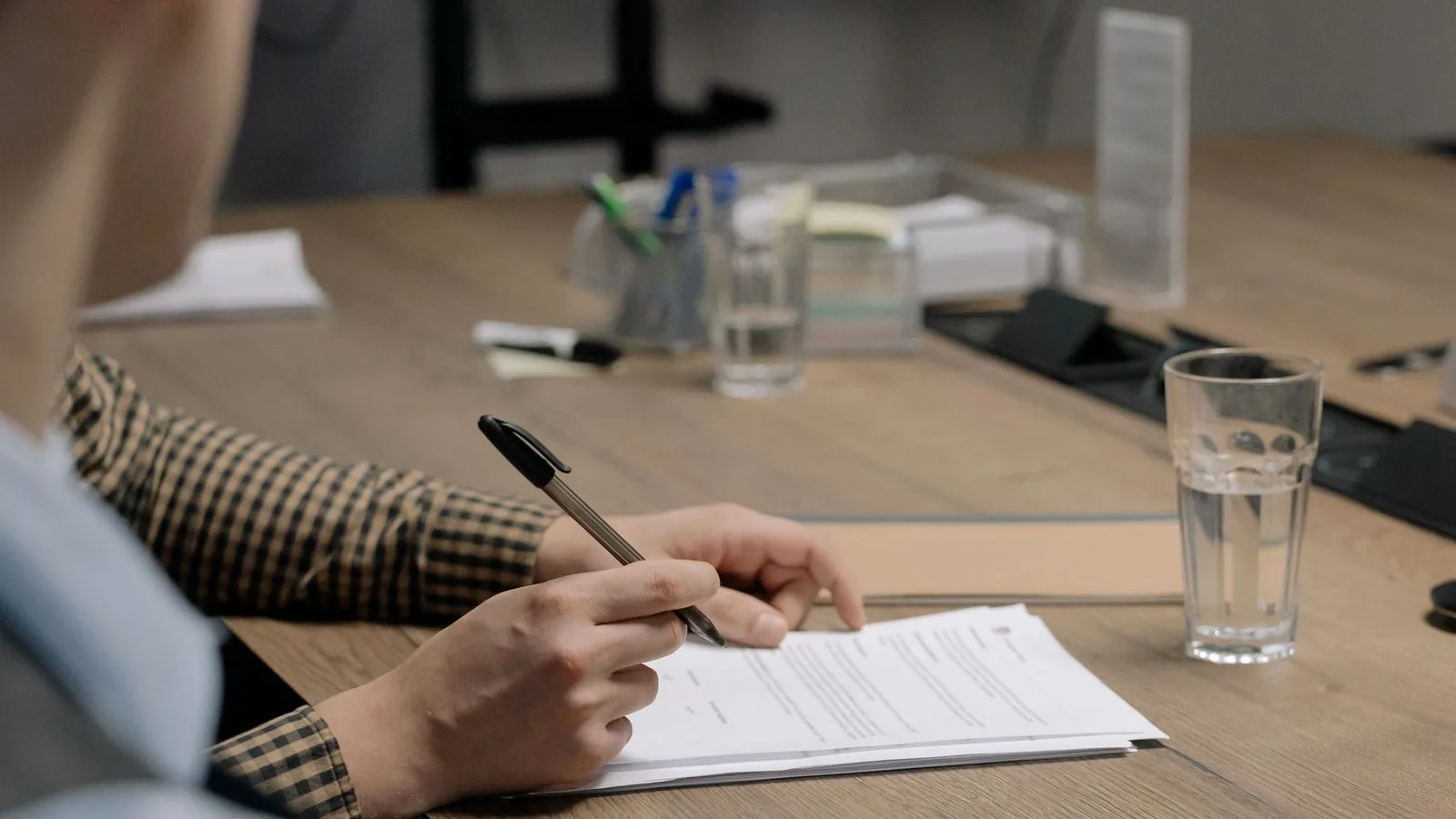 Person in business attire signing a document at a wooden table in an office setting.