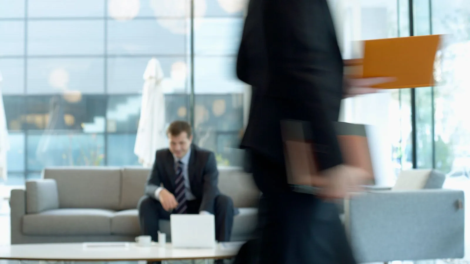 Businessman sitting in modern office lounge using a laptop while colleague walks past, creating dynamic blur.