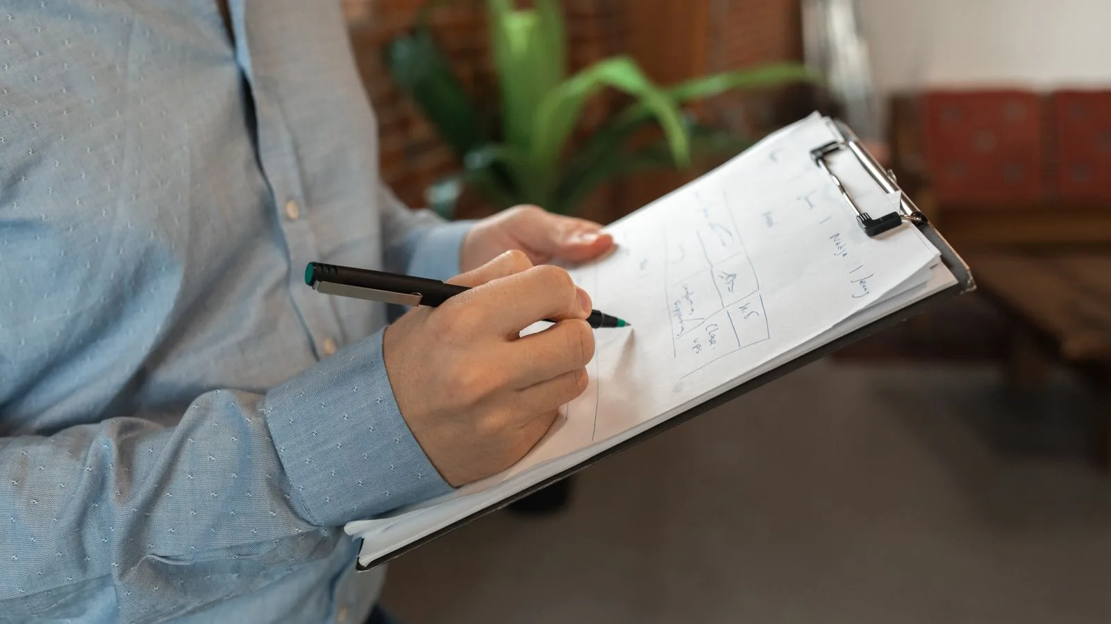 A person in blue shirt writes with a pen on a clipboard indoors, showcasing focus and productivity.