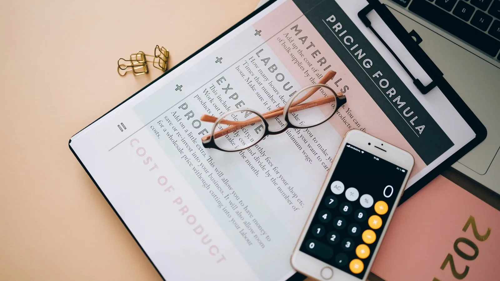 Overhead view of business tools including a phone calculator, pricing formula document, and eyeglasses on a desk.