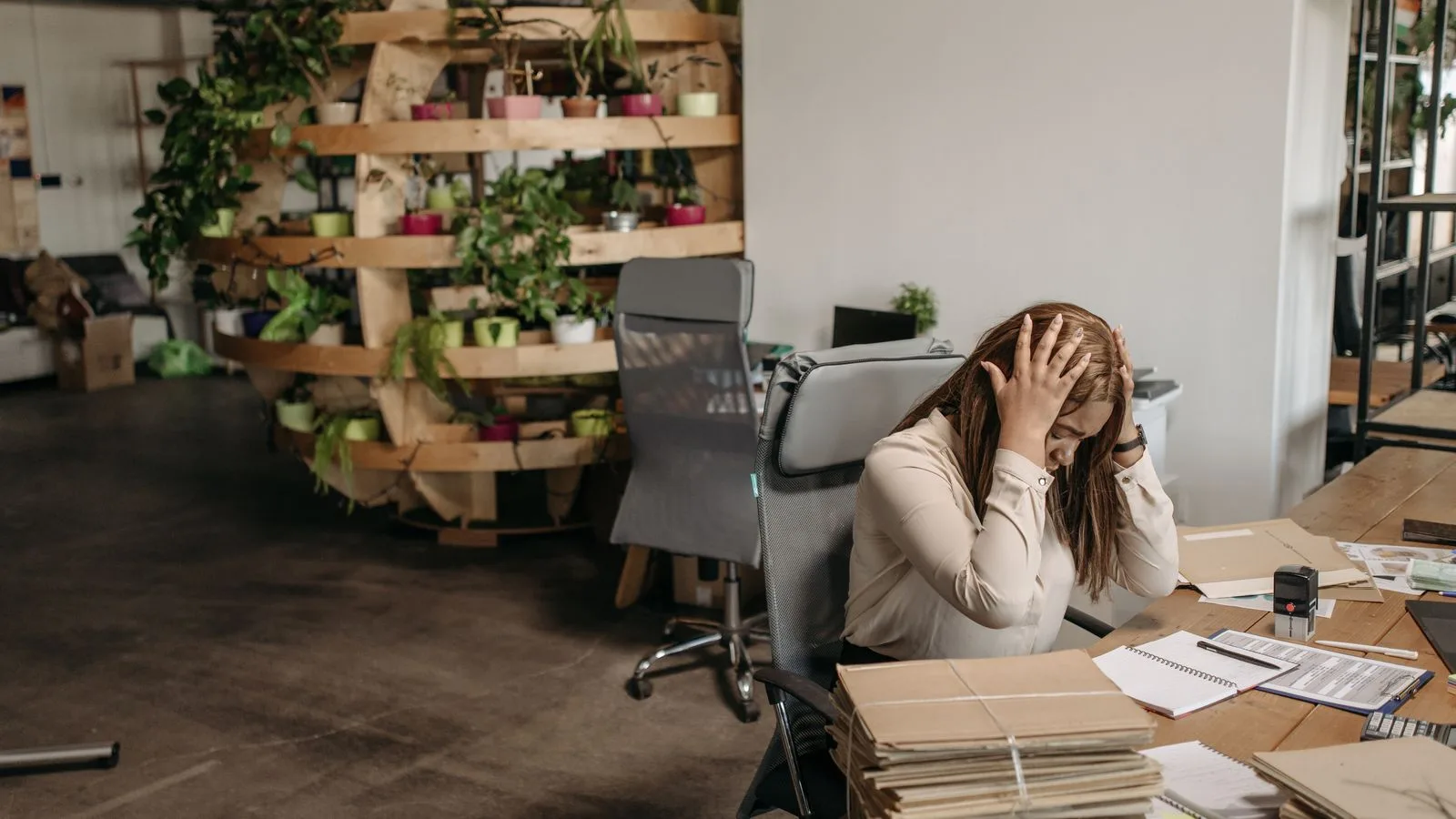 A stressed woman sits overwhelmed at her desk, surrounded by paperwork in a modern office setting.