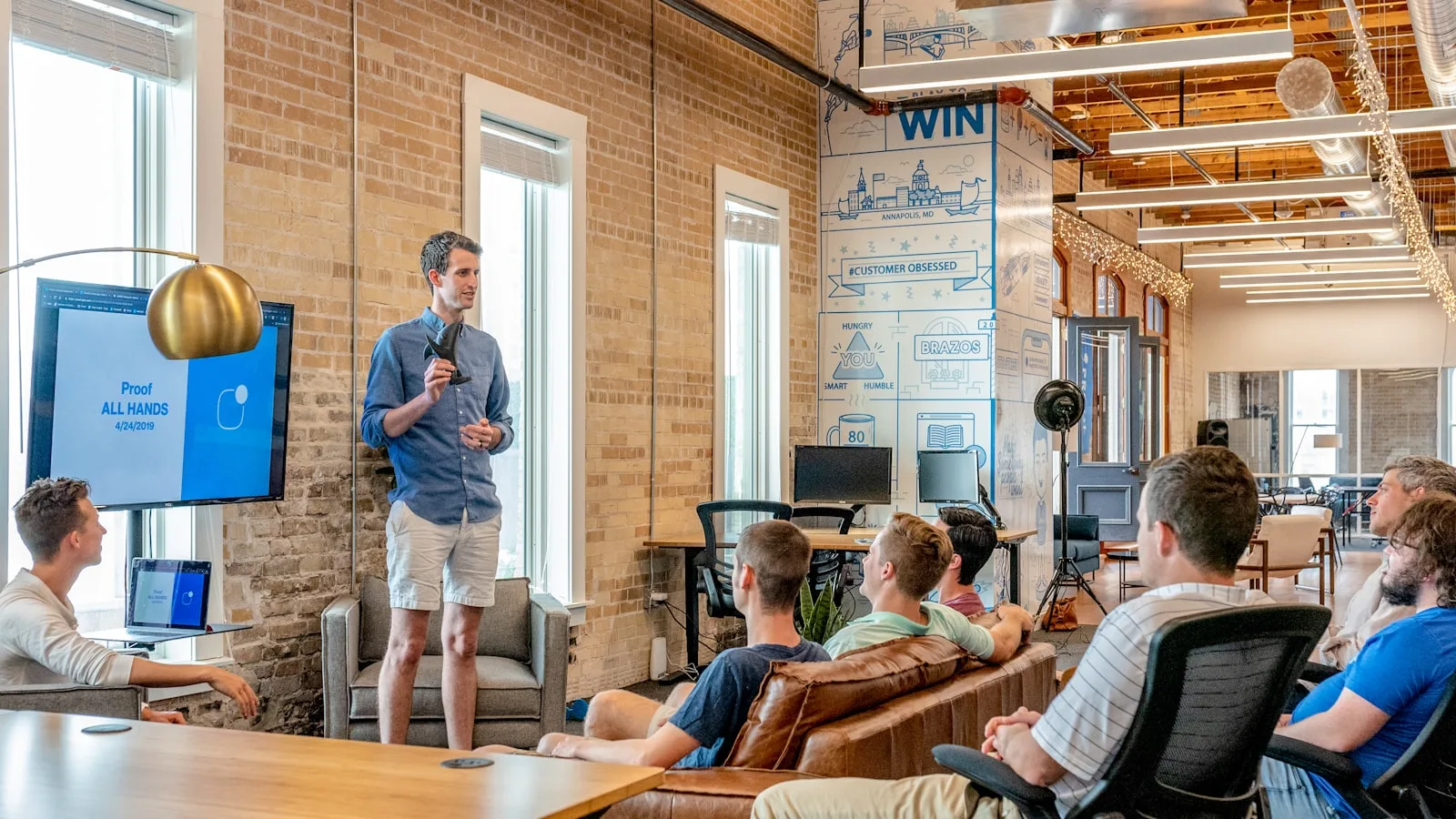 A team lead presenting to colleagues seated on couches in an open office