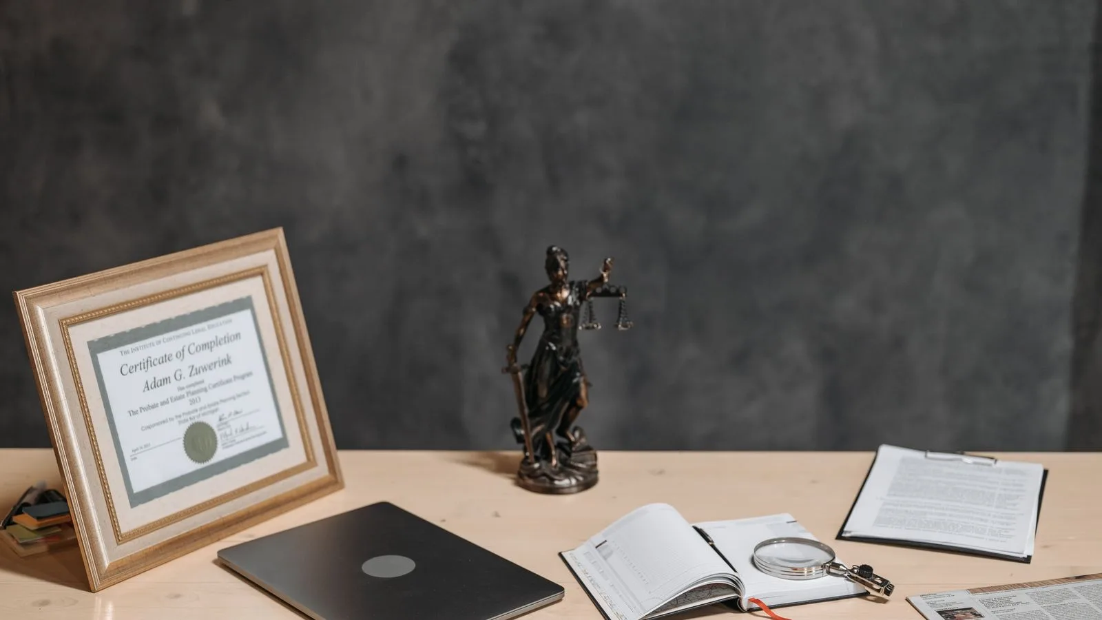 A lawyer's desk featuring a justice statue, diploma, laptop, magnifying glass, and documents.