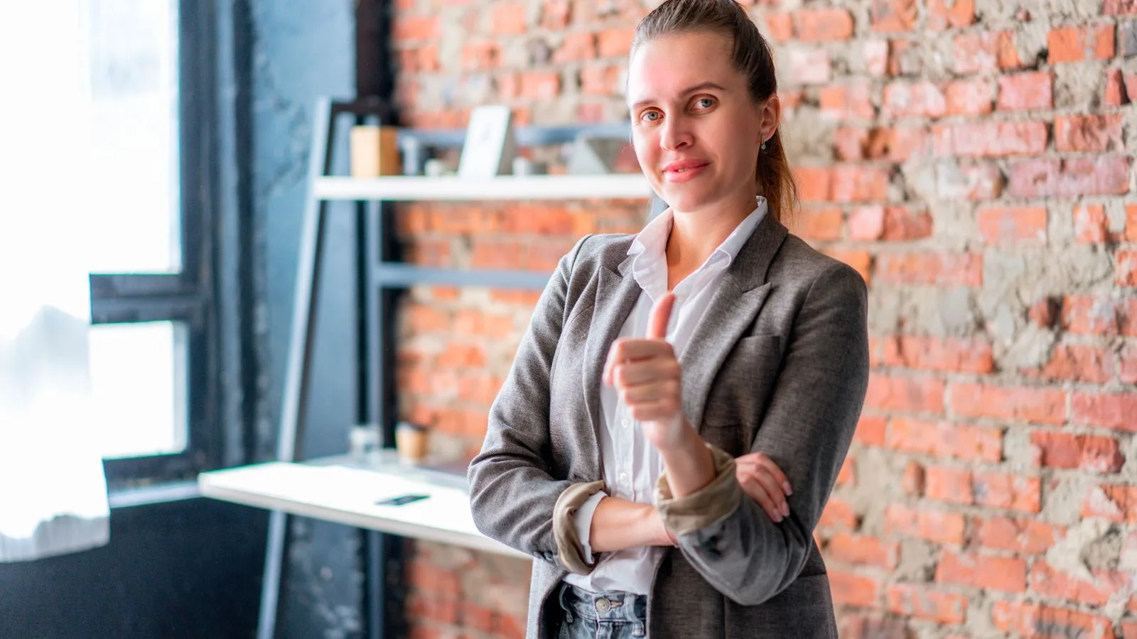 Professional businesswoman giving thumbs up in a stylish office with brick walls.